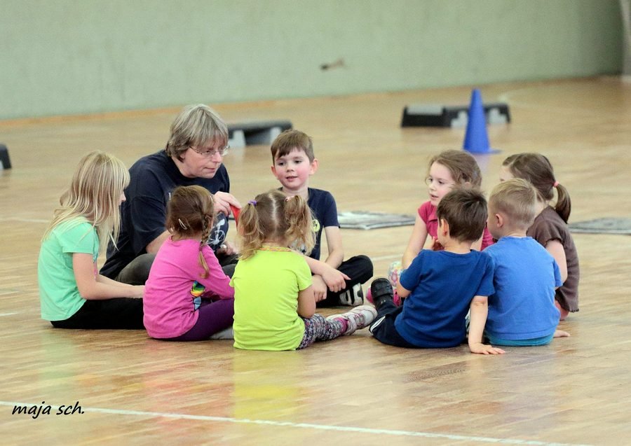 Die Übungsleiterin sitzt mit den Kindern im Kreis auf dem Hallenboden.