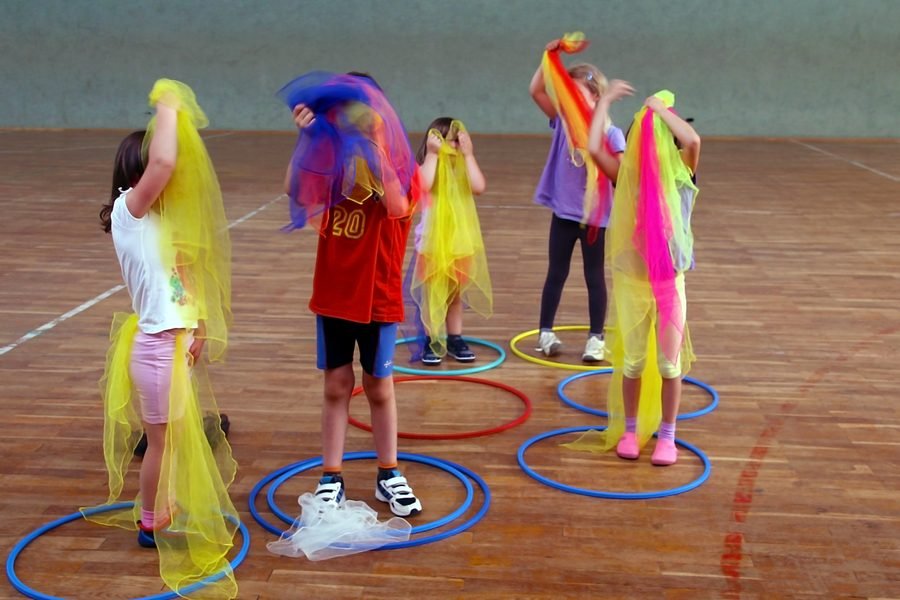 Kinder tanzen mit bunten Tüchern zwischen Reifen in der Turnhalle.
